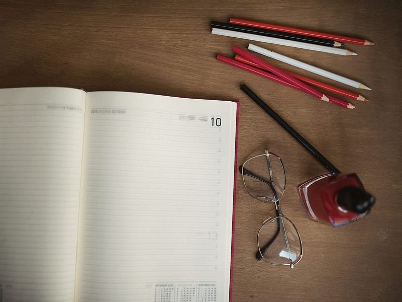 An open weekly planner with coloured pencils and glasses on a wooden desk